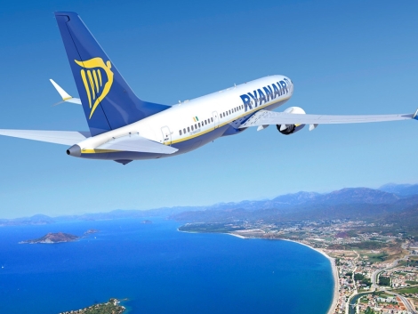 Lietadlo Boeing 737 v modro-žltých farbách Ryanairu stúpa nad morské pobrežie s plážou a mestskou zástavbou