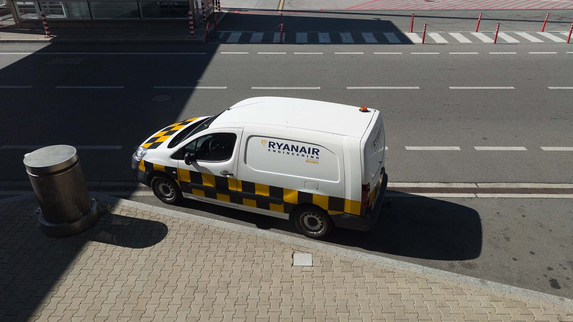 Servisná dodávka s logom Ryanair Engineering stojí pri skle terminálu, v pozadí pracovníci handlingu v reflexných vestách a lietadlo na stojisku