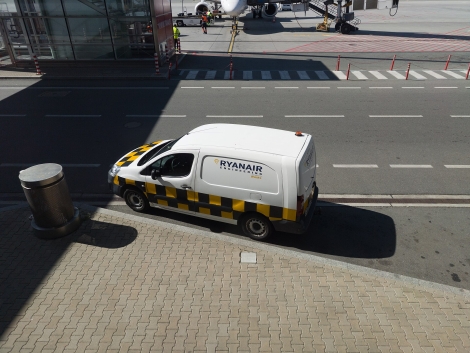 Servisná dodávka s logom Ryanair Engineering stojí pri skle terminálu, v pozadí pracovníci handlingu v reflexných vestách a lietadlo na stojisku